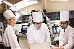 A mixed race group of chefs working with a laptop computer in a commercial kitchen.