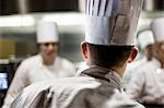 A closeup view from behind of a chef  wearing a toque hat in a commercial kitchen.