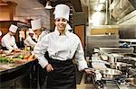 A portrait of a young black female chef in a commercial kitchen with her crew working behind her.