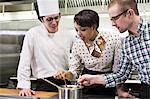 A Caucasian male chef teaching a cooking class for a mixed race grope of students in a commercial kitchen,