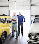 A portrait of a Caucasian senior car mechanic and his son in their classic car repair shop.