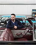 A portrait of a Caucasian male leaving on the side of his classic car convertible in a repair shop.