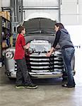 A Caucasian female car mechanic talks to her young son about a car engine in a classic car repair shop.