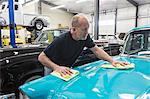 A Caucasian senior male car mechanic applying wax to the hood of an old sedan in his classic car repair shop.