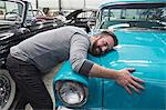 A Caucasian male hugging the hood of his old sedan in a classic car repair shop.