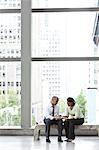 A black businesswoman and a middle eastern businessman sitting on a bench in a convention centre lobby and checking a text on a cell phone.