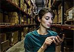 A Caucasian female warehouse worker checking her phone for texts while standing in a distribution warehouse.