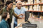 A group of three mixed race warehouse workers checking inventory on a computer in a large distribution warehouse.