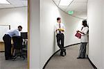 Two Asian businesspeople talking in a hallway near a black businessman working alone in his office.