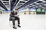 A Caucasian businessman texting while sitting in a chair in the middle of a convention centre arena.