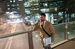 Businessman talking on smart phone, walking on urban pedestrian bridge at night