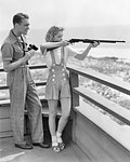 1940s YOUNG COUPLE SHOOTING 22 CALIBER PUMP RIFLE ATTRACTIVE BLOND WOMAN AIMING AND FIRING SMILING MAN WITH BINOCULARS COACHING