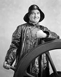 1930s SMILING MAN SAILOR WEARING OILSKIN SOU'WESTER HAT AND RAIN COAT LOOKING AT CAMERA STANDING WATCH AT SHIP'S WHEEL