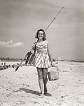 1940s SMILING YOUNG WOMAN WALKING ON BEACH LOOKING AT CAMERA WEARING TWO PIECE BATHING SUIT SKIRT CARRYING SURF FISHING GEAR