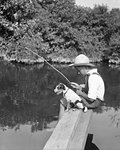 1930s BOY WEARING STRAW HAT SITTING ON WOODEN PLANK WITH DOG FISHING IN POND