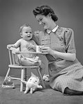 1940s SMILING MOTHER SUPPORTING ONE YEAR OLD BABY SON SITTING IN CHAIR AMONG VARIOUS TOYS