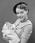 1950s PORTRAIT SMILING YOUNG WOMAN MOTHER WEARING BERET HAT LOOKING AT CAMERA HOLDING INFANT BABY SON IN ARMS