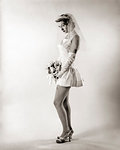 1950s SEXY SEDUCTIVE WOMAN MODEL BRIDE LOOKING AT CAMERA WEARING VERY SHORT WEDDING DRESS VEIL AND HIGH HEELS HOLDING BOUQUET