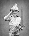 1930s SMILING LITTLE BOY WEARING FOLDED NEWSPAPER FORE AND AFT COCKED HAT HOLDING WOODEN SWORD HAND SALUTING LOOKING AT CAMERA