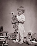 1930s YOUNG BLOND BABY BOY STANDING WEARING WOOL SOCK FOOTED DR. DENTON SLEEPWARE HOLDING A FAVORITE JESTER DOLL AMID OTHER TOYS