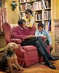 1970s AFRICAN AMERICAN FATHER READING TO SON AND DAUGHTER IN LIVING ROOM WITH AIREDALE DOG SITTING BY CHAIR