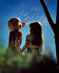 1970s 1980s BLOND BOY AND BRUNETTE GIRL HOLDING HANDS SITTING TOGETHER UNDER TREE BOY BLOWING BUBBLES INTO BLUE SKY