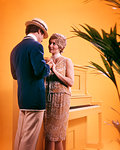 1970s COUPLE MAN WOMAN WEARING 1920s ERA JAZZ AGE CLOTHES HOLDING HANDS STANDING BY PAINTED YELLOW UPRIGHT PIANO