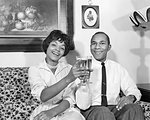 1960s SMILING AFRICAN AMERICAN HUSBAND AND WIFE SITTING IN LIVING ROOM LOOKING AT CAMERA TOASTING WITH RAISED GLASSES OF BEER