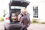 Young man in trilby removing luggage from car boot outside hotel, rear view