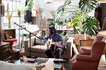 Young male musician recording music, playing guitar and singing into microphone in apartment