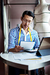 Japanese male fashion designer working in his studio, sitting at table, looking at digital tablet.