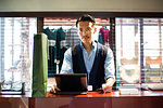 Japanese salesman with moustache wearing glasses standing at counter in clothing store, looking at digital tablet.