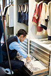 Japanese salesman with moustache wearing glasses kneeling in front of open drawer in clothing store.