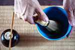 High angle close up of person using bamboo whisk to prepare Matcha tea in a blue bowl during tea ceremony.