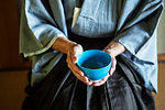 High angle close up of Japanese man wearing traditional kimono kneeling on floor holding blue tea bowl during tea ceremony.