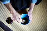 High angle close up of Japanese man wearing traditional kimono kneeling on floor using bamboo whisk to prepare Matcha tea in a blue bowl during tea ceremony.
