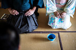 High angle view of Japanese man and woman wearing traditional white kimono with blue floral pattern kneeling on floor during tea ceremony, holding blue tea bowl.