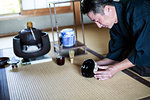 Japanese man wearing traditional kimono kneeling on floor, holding tea bowl, during tea ceremony.