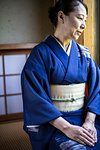 Japanese woman wearing traditional bright blue kimono with cream coloured obi kneeling on floor in traditional Japanese house.