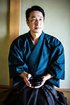 Japanese man wearing kimono kneeling on floor, holding tea bowl during tea ceremony.