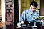 Japanese man wearing kimono sitting on floor in traditional Japanese house, using calculator and digital tablet.