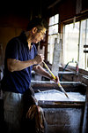 Japanese man stirring a vat of liquid in a workshop with a stick, making traditional Washi paper.