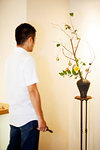 Japanese man standing in flower gallery, looking at Ikebana arrangement in brown vase.