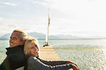 Mature couple on jetty, sitting back to back, smiling