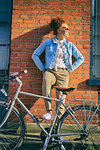 Young woman with bicycle wearing sunglasses leaning against brick wall, hands on hips looking away
