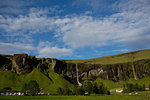 Remote village by waterfall flowing over lush green mountain range, Iceland