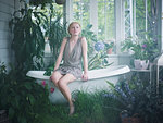 Young woman sitting on edge of bathtub, in bathroom filled with plants