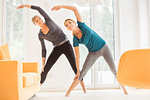 Two mature women bending over sideways whilst exercising in living room