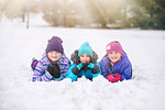 Friends wearing knit hats lying on front side by side in snow looking at camera smiling