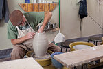 Potter wearing flat cap sitting at pottery wheel shaping clay vase